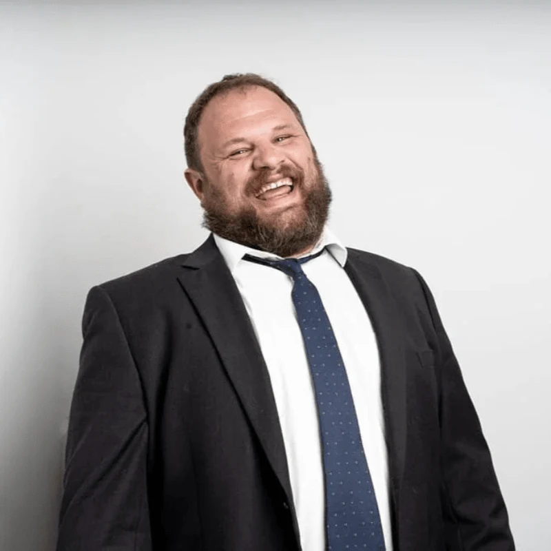 Comedian Sam Miller wearing suit jacket and tie, laughing.